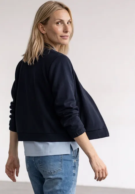 Seitlich stehende Frau mit blonder Mähne, dunkelblauer Jacke und Jeans, schaut über die Schulter.