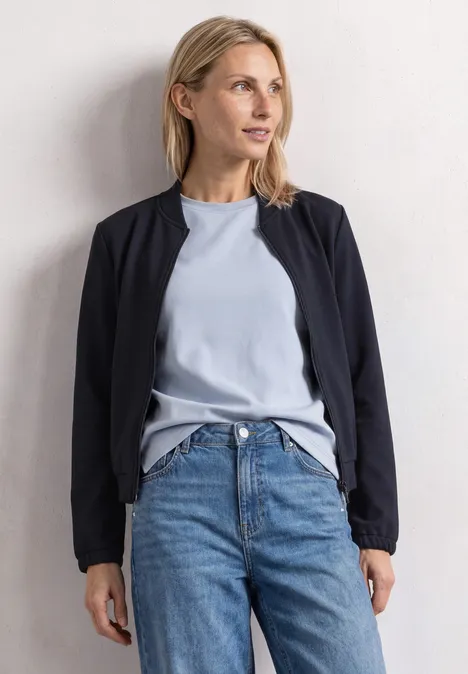 Frau in Jeans, hellblauem T-Shirt und schwarzer Jacke lehnt entspannt an weißer Wand.