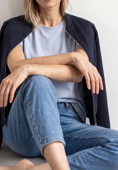 Frau sitzt mit verschränkten Beinen, trägt Jeans, helles T-Shirt und lässig auf den Schultern liegenden Blazer.