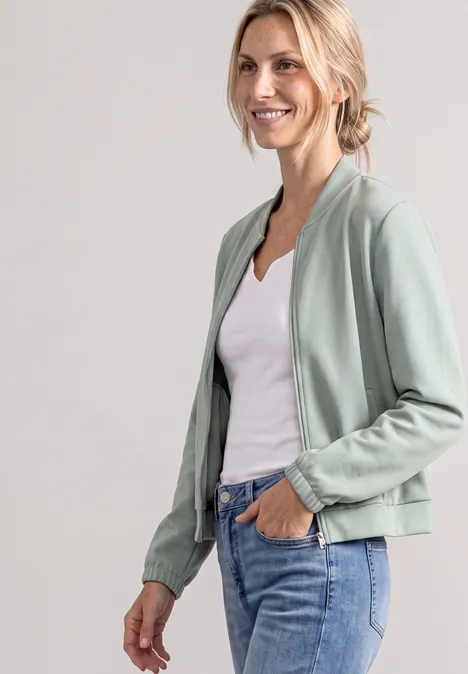Frau trägt hellgrüne Bomberjacke, weißes Top und Jeans, lächelt entspannt