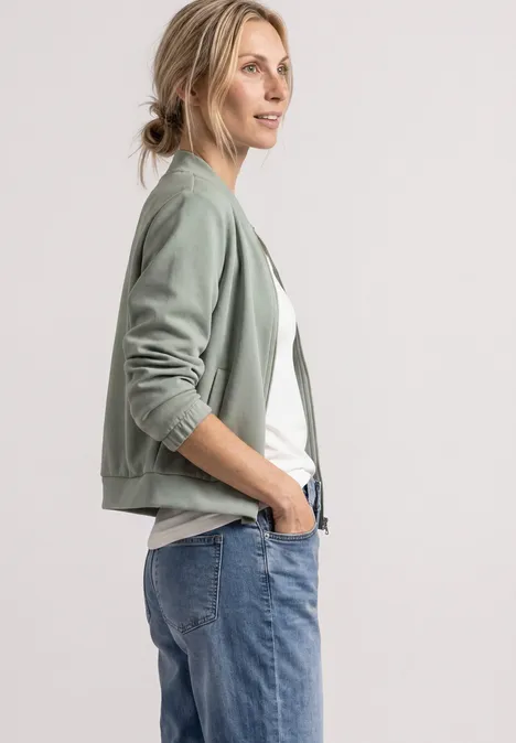 Frau trägt grünen Blouson, weißes Shirt und blaue Jeans, sie steht seitlich und lächelt leicht.