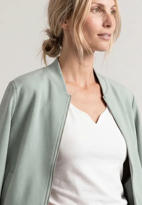 Blonde Frau in weißem Shirt und mintgrünem Blouson, seitlich in die Ferne blickend.
