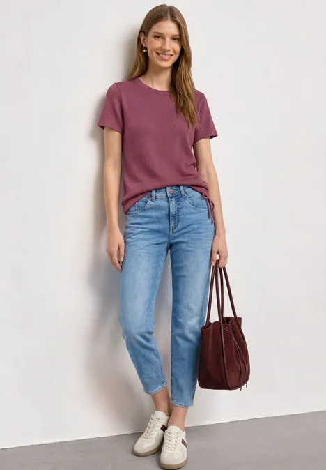 Basic T-Shirt im Strick-Look mit Rundhals dark clouded blush Frau lehnt an Wand, trägt bordeauxrotes T-Shirt, blaue Jeans und weiße Sneakers, hält rote Tasche.