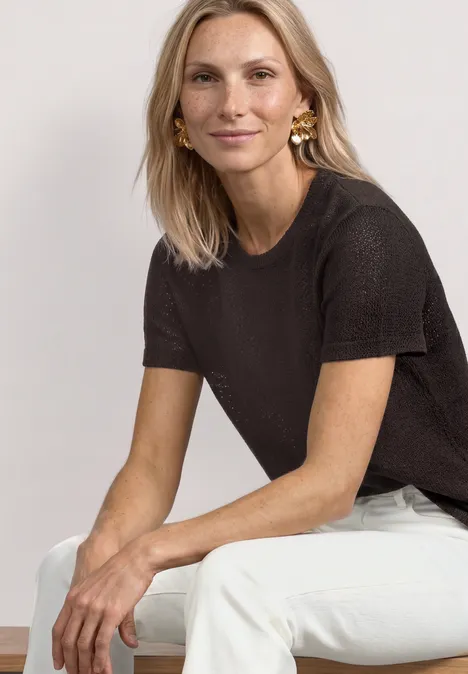 Basic T-Shirt im Strick-Look mit Rundhals black coffee Eine Frau sitzt entspannt seitlich auf einer Bank, trägt ein schwarzes T-Shirt und weiße Hose.