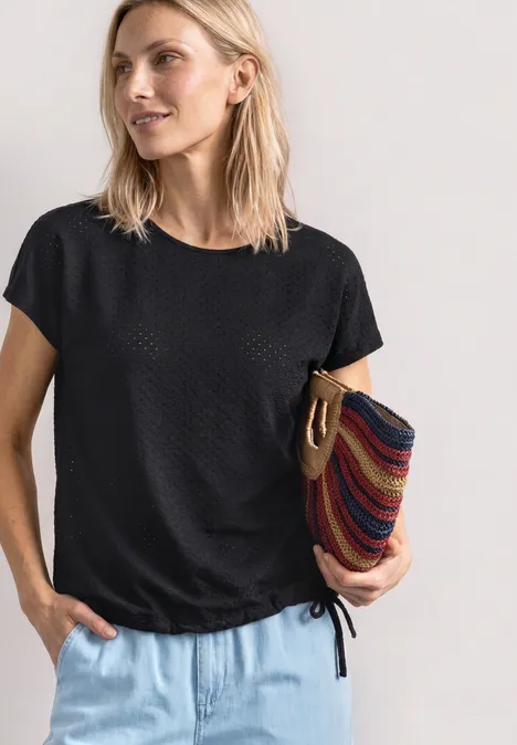 T-Shirt aus Spitze mit Rundhals Black Frau trägt schwarzes Kurzarmshirt, helle Jeans und hält bunte gestreifte Tasche.