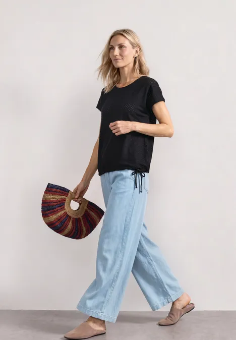 T-Shirt aus Spitze mit Rundhals Black Frau in schwarzem T-Shirt, hellblauer weiter Hose und Sandalen, die eine fächerförmige Tasche hält.