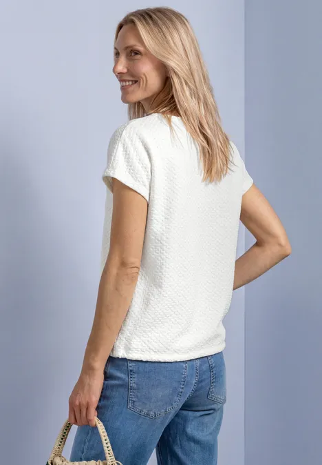 T-Shirt aus Spitze mit Rundhals off white Eine blonde Frau steht seitlich, trägt ein weißes Shirt und Jeans, hält eine Strohtasche und lächelt.