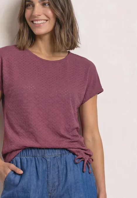 T-Shirt aus Spitze mit Rundhals dark clouded blush Frau in lockerem weinrotem Shirt und blauen Freizeithosen lächelt vor hellem Hintergrund.