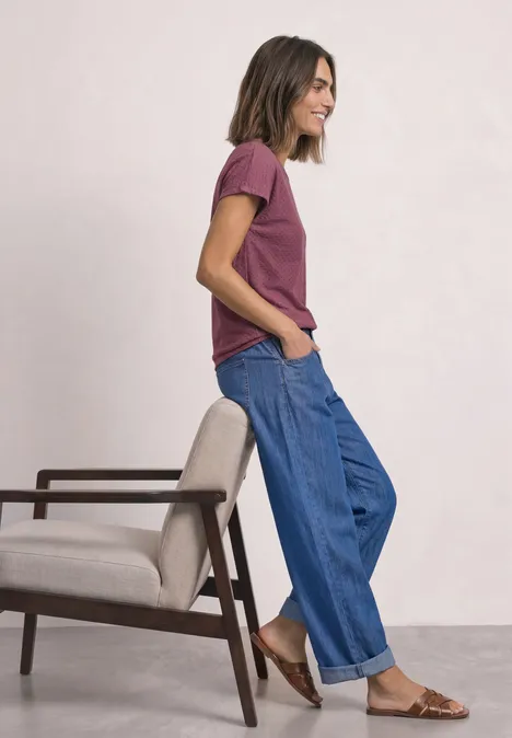T-Shirt aus Spitze mit Rundhals dark clouded blush Frau in lässigem Outfit mit T-Shirt, weiten Jeans und Sandalen neben einem Stuhl, seitlich stehend.