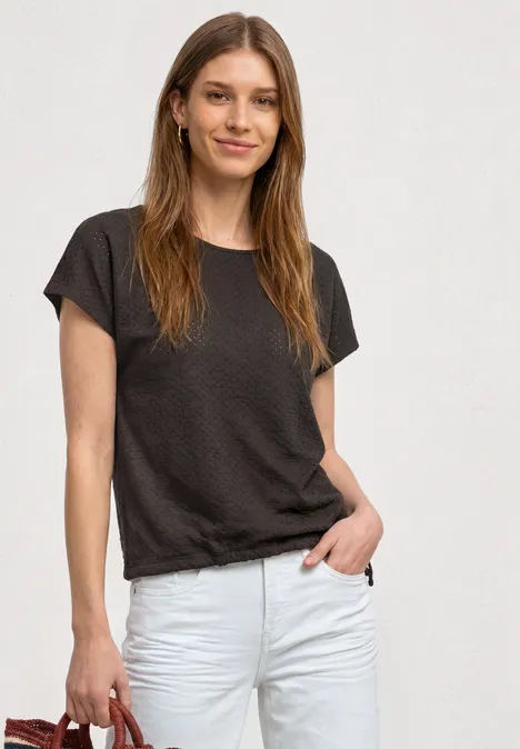 T-Shirt aus Spitze mit Rundhals black coffee Junge Frau im dunklen T-Shirt und weißen Jeans, lächelnd vor hellem Hintergrund.