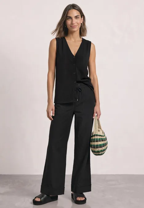 Top mit V-Neck und dekorativer Knopfleiste Black Frau trägt schwarzes ärmelloses Top und weite Hose, hält Korbtasche, trägt Sandalen.