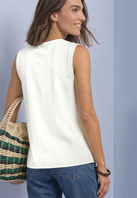 Top met V-hals en decoratieve knoopsluiting off white Vrouw in mouwloos wit shirt en spijkerbroek, met gevlochten handtas, lacht en kijkt opzij.