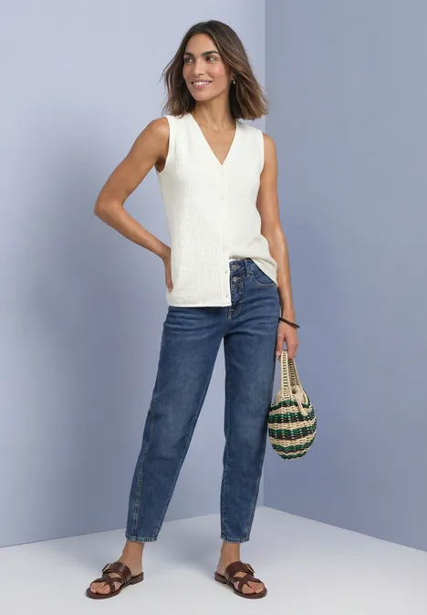 Top met V-hals en decoratieve knoopsluiting off white Vrouw in witte mouwloze top, spijkerbroek en sandalen, met rieten tas voor paarse achtergrond.
