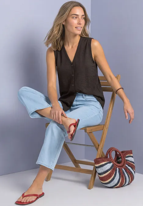 Top mit V-Neck und dekorativer Knopfleiste black coffee Frau in schwarzem Top, hellen Jeans und roten Sandalen sitzt lässig auf einem Holzstuhl, Korbtasche daneben.