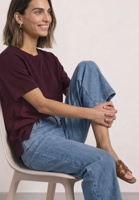 Frau sitzt lächelnd auf einem Stuhl, trägt bordeauxrotes Shirt, blaue Jeans und braune Schuhe.