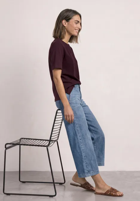 Frau sitzt seitlich auf einem Drahtstuhl, trägt Burgundershirt, weite Jeans und braune Sandalen vor hellem Hintergrund.