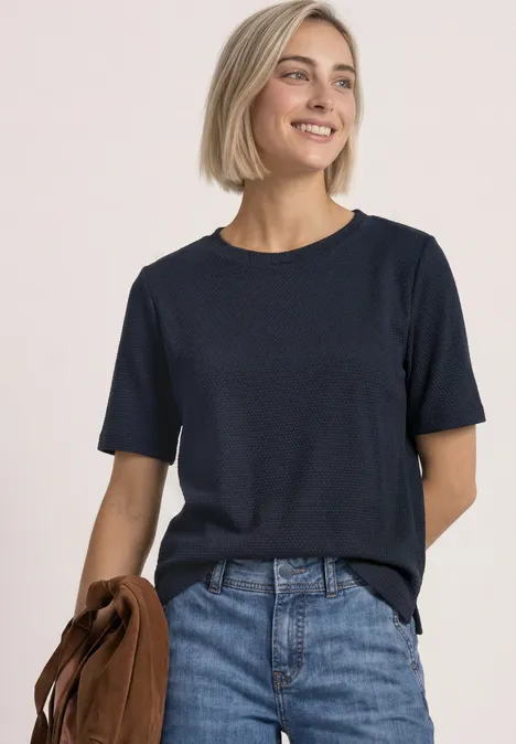 Femme aux cheveux blonds courts portant un t-shirt bleu marine et un jean, souriante, tenant une veste marron.