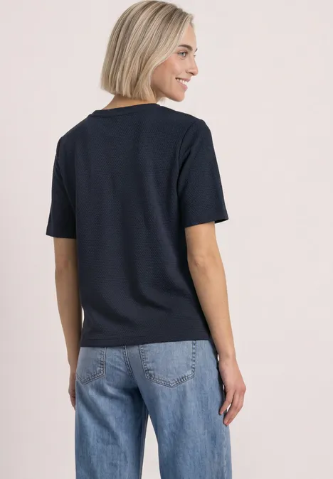 Femme aux cheveux blonds courts porte un T-shirt noir et un jean bleu, vue de dos.