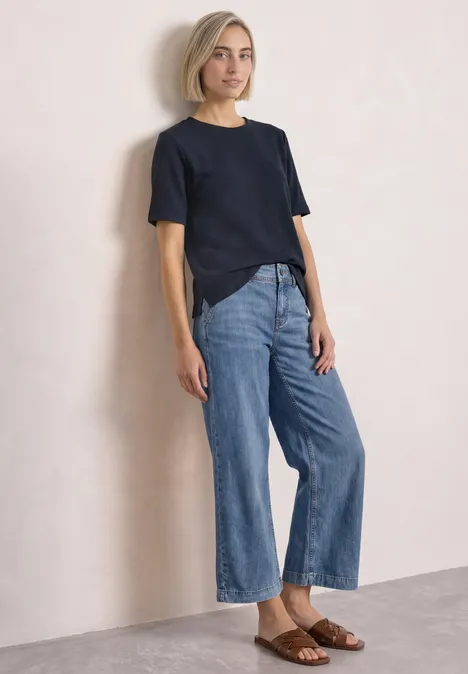 Femme en t-shirt noir, jean large et sandales brunes sur fond clair.