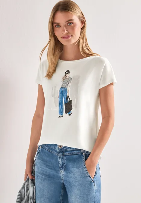 Frau trägt weißes T-Shirt mit Illustration einer Frau in blauer Hose und Jeanshose, Hände in Taschen.