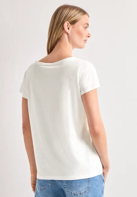Rückansicht einer Frau in weißem T-Shirt und Jeans mit Händen in den Taschen vor heller Wand.