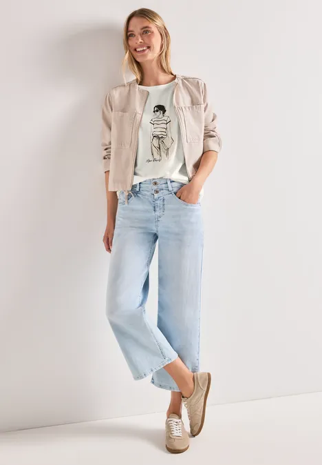 Frau trägt helle Jeans, weißes Grafik-T-Shirt, beigen Blazer und Slipper, steht lässig.