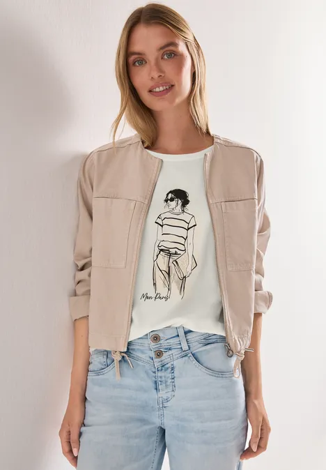 Frau in blauen Jeans, weißem T-Shirt mit Skizze und heller Bomberjacke vor weißem Hintergrund.