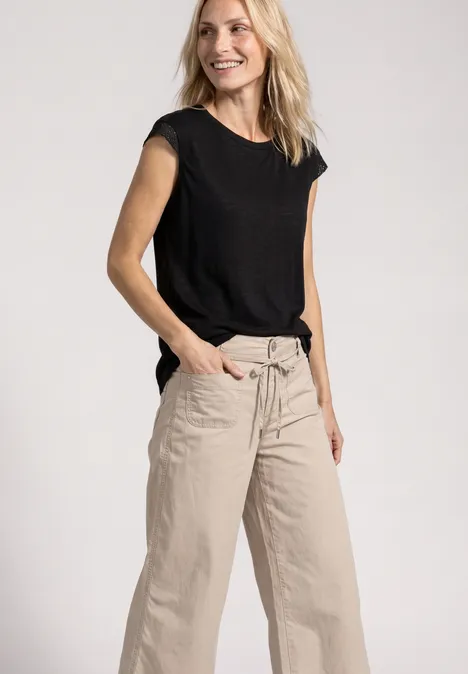 Eine Frau trägt ein schwarzes T-Shirt und beige weite Hosen, lächelt und hat die Hände in den Taschen.