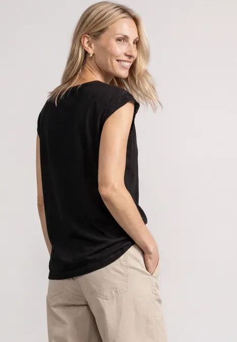 Seitliche Ansicht einer Frau in schwarzem Shirt und beiger Hose, lächelnd mit den Händen in den Taschen.