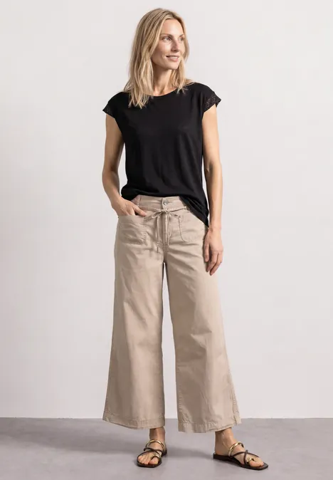 Schlanke Frau im schwarzen T-Shirt, beigen Culotte-Hosen und Sandalen, steht lässig mit Händen in den Taschen.