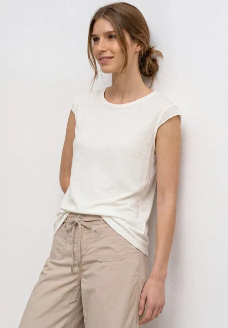 Frau lehnt an weißer Wand, trägt weißes T-Shirt, beige Hose und locker gesteckte Haare.