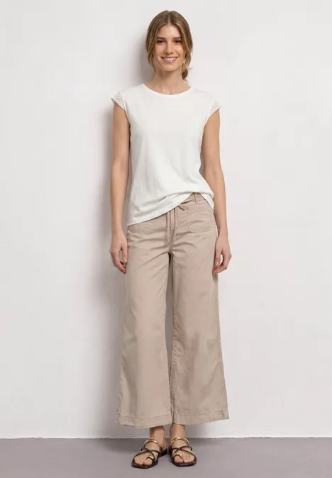 Junge Frau in weißem T-Shirt, beigen Culotte-Hosen und Sandalen vor neutralem Hintergrund stehend.