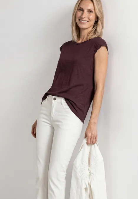 Frau in weißer Jeans und bordeauxrotem Shirt hält eine weiße Jacke vor neutralem Hintergrund.