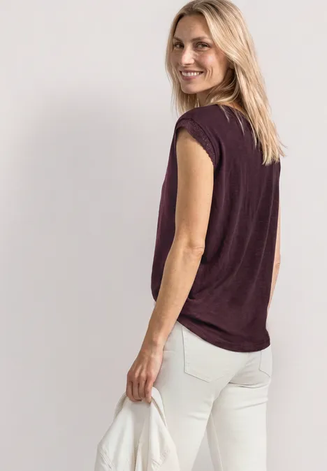 Frau mit blondem Haar trägt ein ärmelloses violettes Top und helle Hose, seitliche Ansicht.