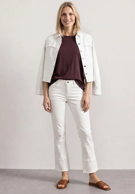 Frau in weißer Jeansjacke, auberginefarbenem Shirt, weißen Jeans und braunen Sandalen vor neutralem Hintergrund.
