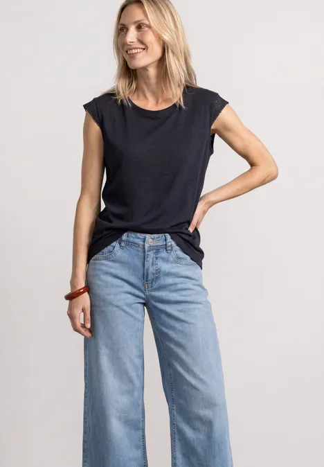 Frau in schwarzem T-Shirt und hellblauen, weiten Jeans steht vor neutralem Hintergrund.