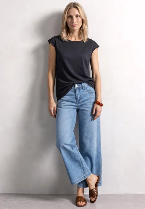 Frau in schwarzem T-Shirt und weiten Jeans steht vor weißer Wand.
