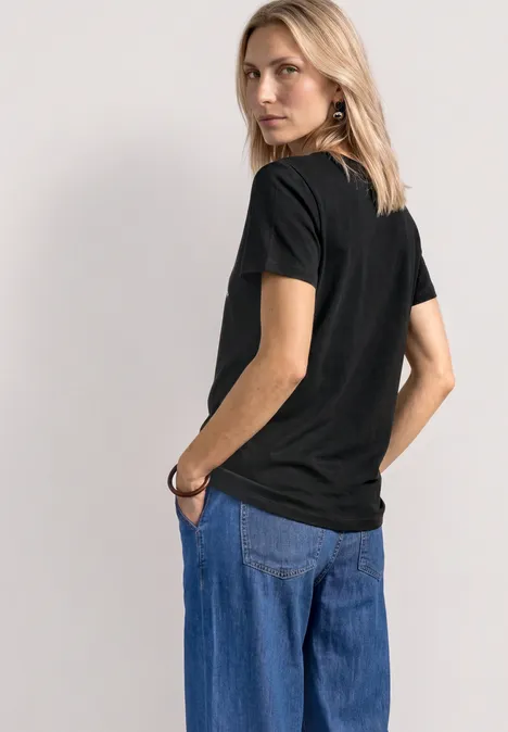 T-Shirt mit Rundhals und Frontprint Black Frau in schwarzem T-Shirt und blauen Jeans, Rückenansicht, eine Hand in der Tasche.