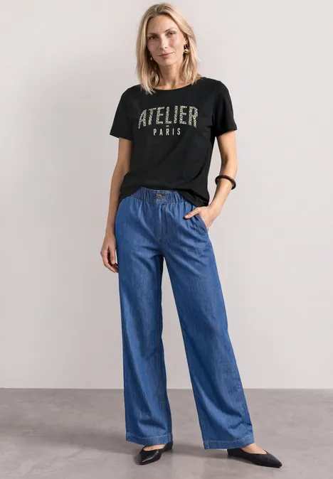 T-Shirt mit Rundhals und Frontprint Black Frau trägt schwarzes T-Shirt mit 'ATELIER PARIS' und weite blaue Jeans, schwarze Schuhe.