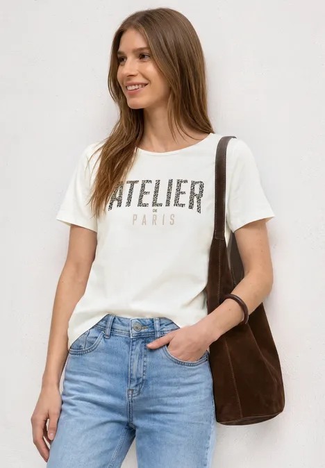 Frau in weißem T-Shirt 'ATELIER PARIS', Jeans, brauner Tasche über der Schulter, vor neutralem Hintergrund.