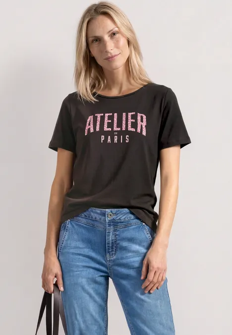 Frau trägt schwarzes T-Shirt mit 'ATELIER PARIS', blaue Jeans und schwarze Tasche.
