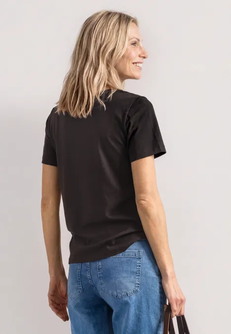 Frau von hinten in schwarzem T-Shirt und blauen Jeans, blondes Haar, vor hellem Hintergrund.