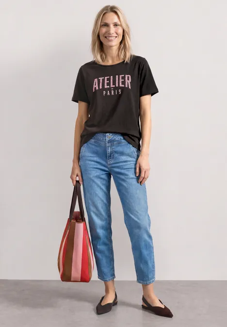 Frau trägt schwarzes T-Shirt mit „Atelier Paris“, blaue Jeans, schwarze Ballerinas und gestreifte Tasche.