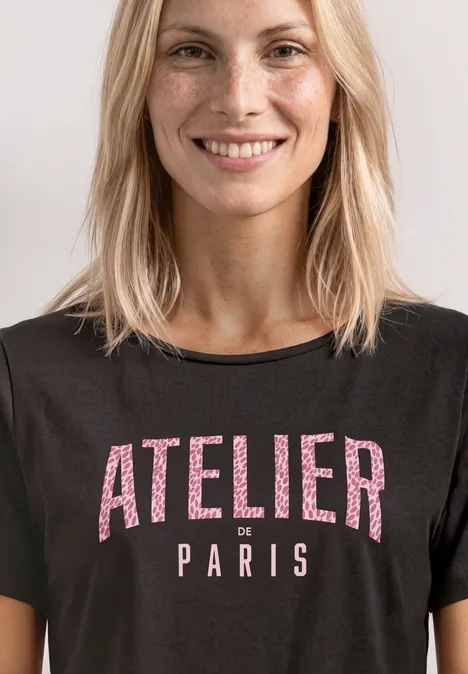 Frau mit blondem Haar trägt schwarzes T-Shirt mit der Aufschrift „ATELIER Paris“.