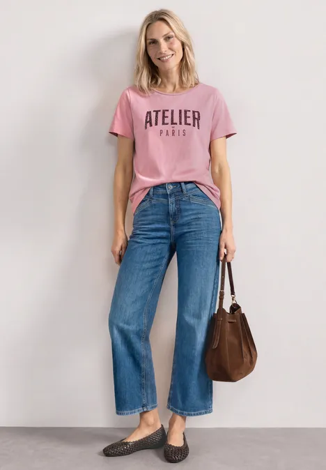 T-Shirt mit Rundhals und Frontprint vintage blush Frau in rosa T-Shirt mit Aufschrift 'ATELIER PARIS', weitem Jeans, Mokassins und brauner Tasche vor weißer Wand.