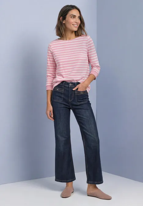Frau mit rosa-weiß gestreiftem Pullover, weiten Jeans und Slippern vor neutralem Hintergrund.