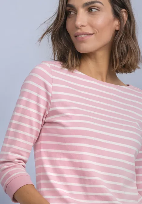Frau mit schulterlangem Braunhaar trägt rosa-weiß gestreiftes Langarmshirt vor hellem Hintergrund.
