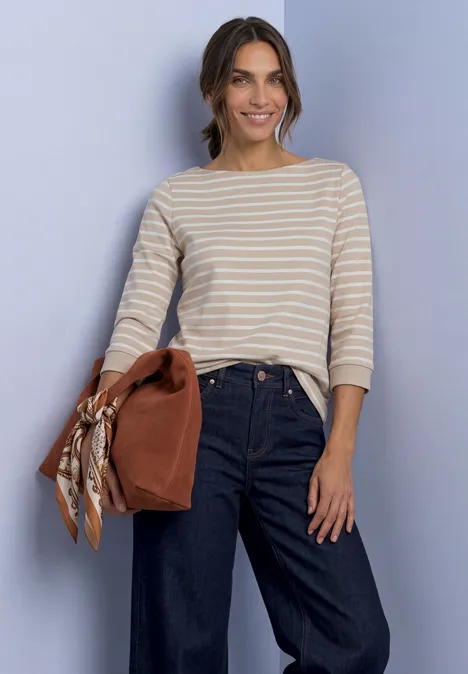 Frau in gestreiftem Shirt und dunklen Jeans hält eine braune Tasche, lächelt locker vor einfachem Hintergrund.