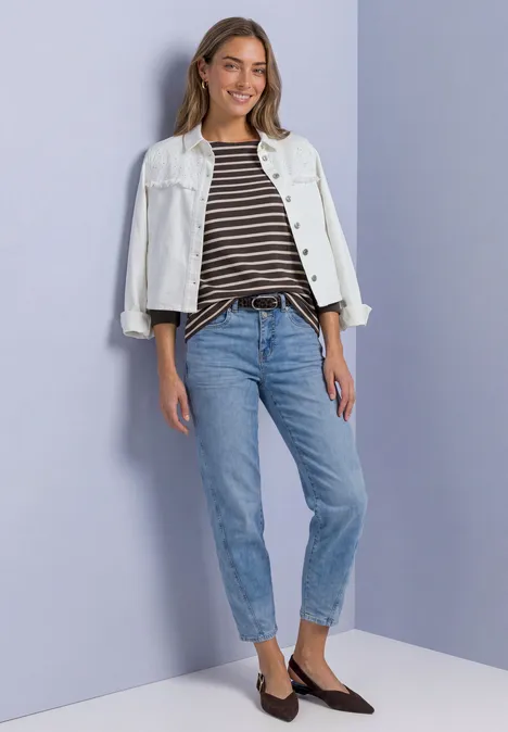 Frau in weißer Jeansjacke, gestreiftem Shirt, blauen Jeans und braunen Slippern vor hellem Hintergrund.