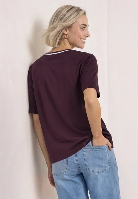 Junge Frau von hinten in lockerem lila T-Shirt mit weißem Kragen und hellblauen Jeans, lässig an Wand gelehnt.